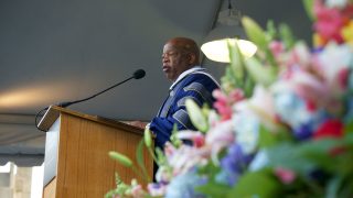 Flowers are in the foreground as John Lewis speaks into the microphone while standing at the lectern in academic regalia.