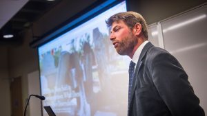 Adam Rothman speaks in front of a projector screen.