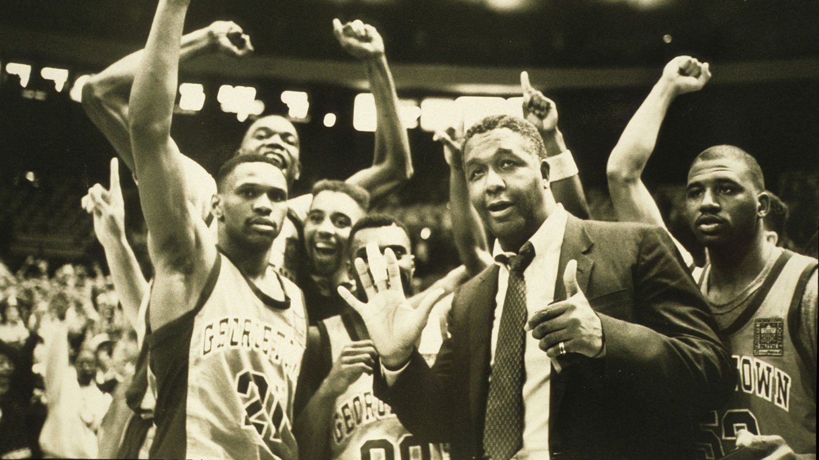 Coach John Thompson Jr. holds up his right palm and left thumb in front of Georgetown basketball players raising their arms in celebration in a faded photo