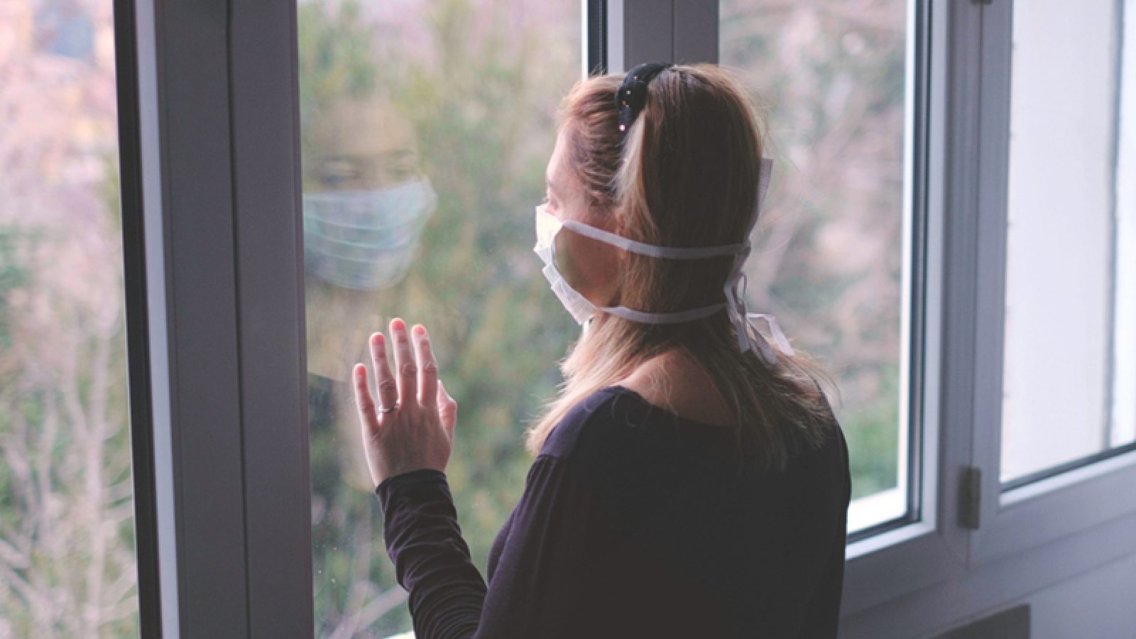 Woman inside wearing mask pressing her hand against a window