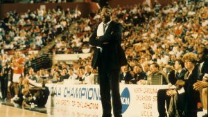 John Thompson Jr. stands courtside with his arms folded and a towel hanging from his shoulder.
