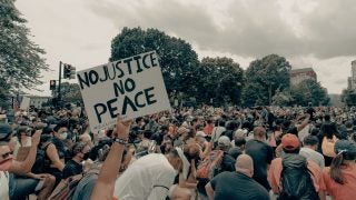 Protestors hold signs while kneeling that read No Justice, No Peace.