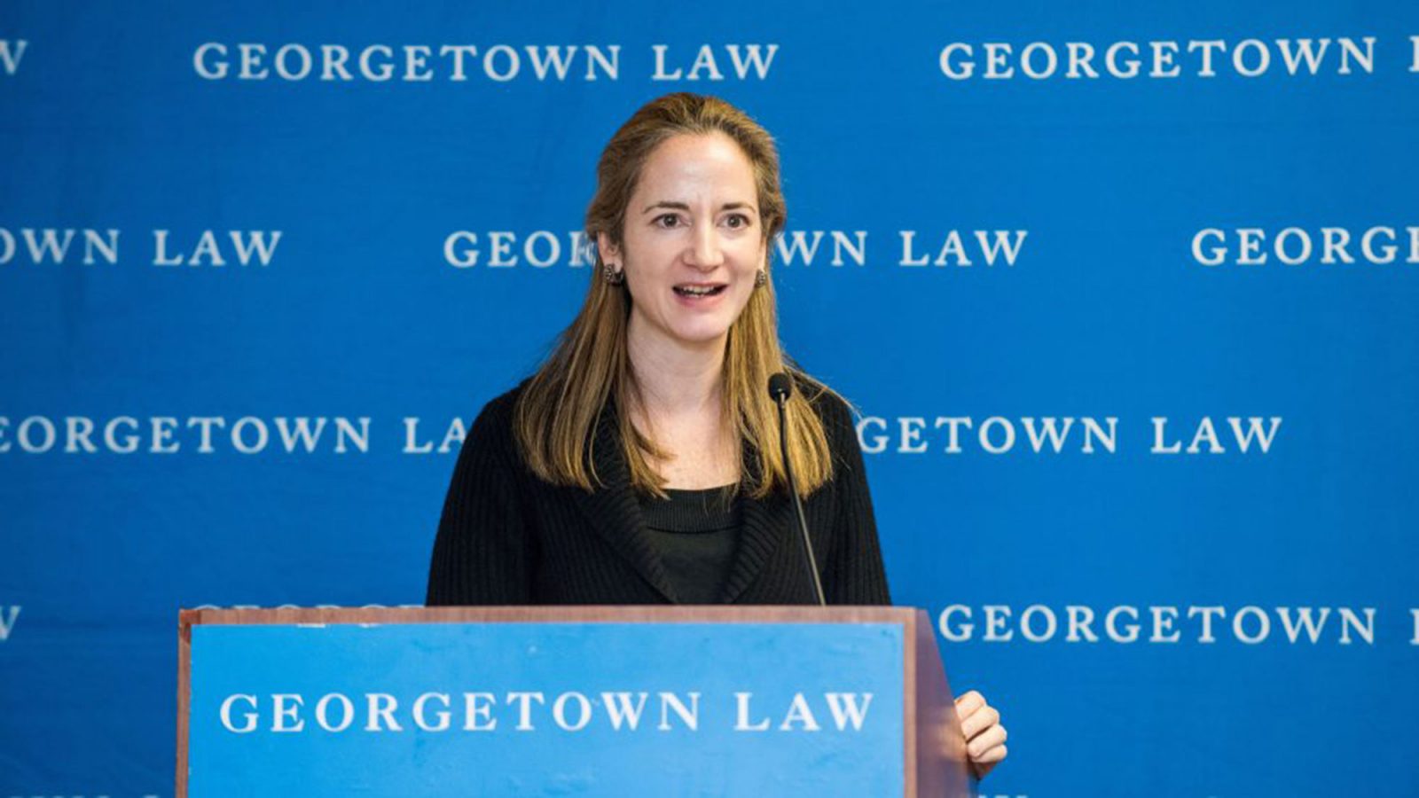 Avril Haines stands at a lectern with a microphone speaking in front of a blue Georgetown Law background.