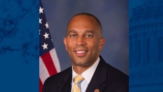 A headshot of Hakeem Jeffries, a McCourt alum in Congress, wearing a black suit with a yellow tie. Behind him is an American flag.