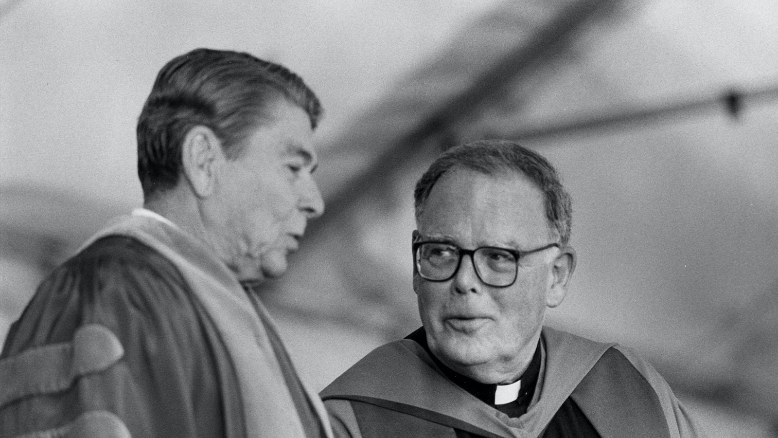 A black and white photo shows of President Ronald Reagan talking to Father Timothy Healy.