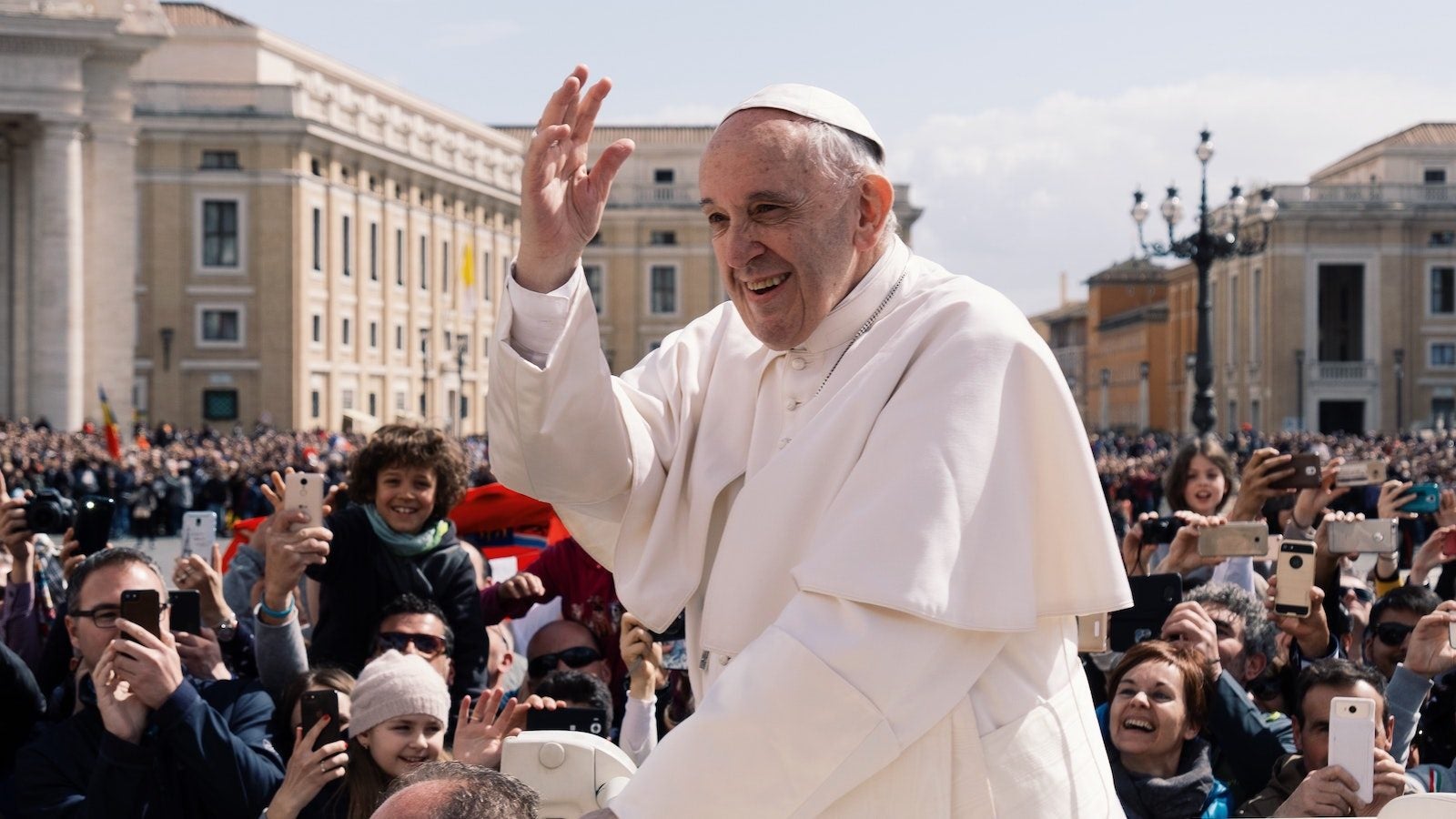 Pope Francis wears white and raises hand in a crowd