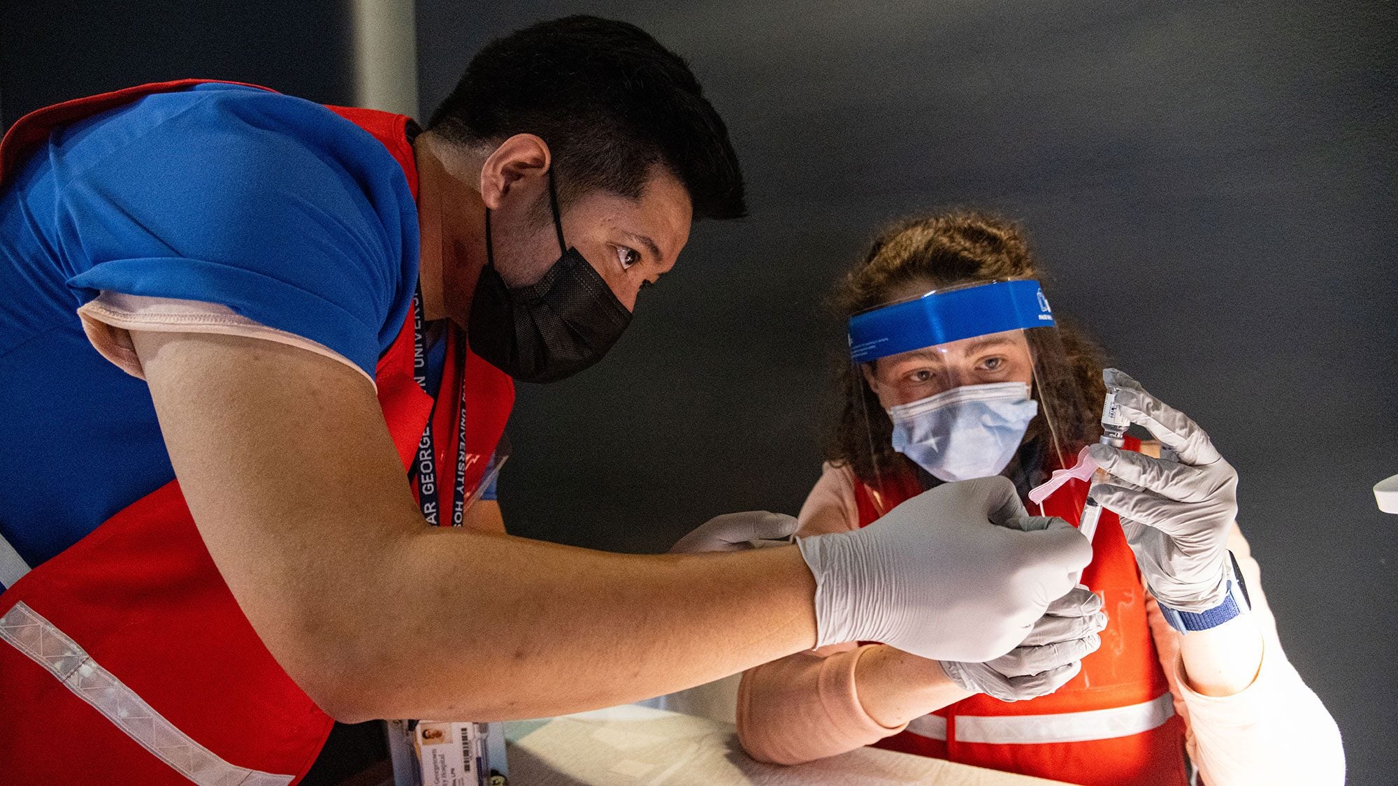 A man and a woman wear masks and a shield whilefilling needles with the vaccine.