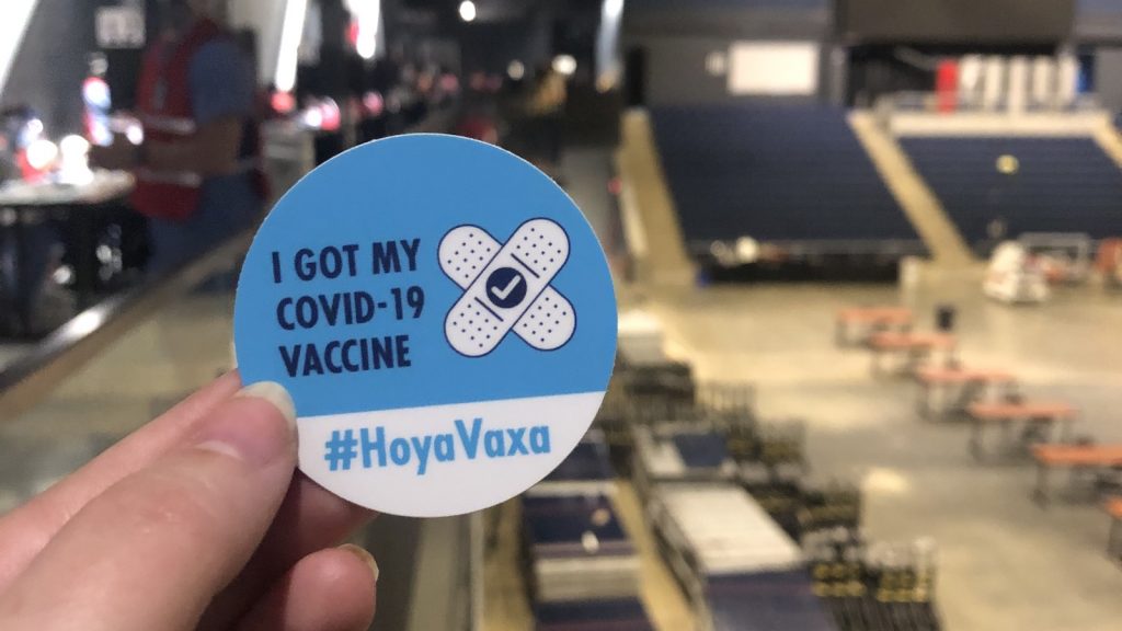 Hand holding a vaccine sticker with text "I got my COVID-19 vaccine #HoyaVaxa"