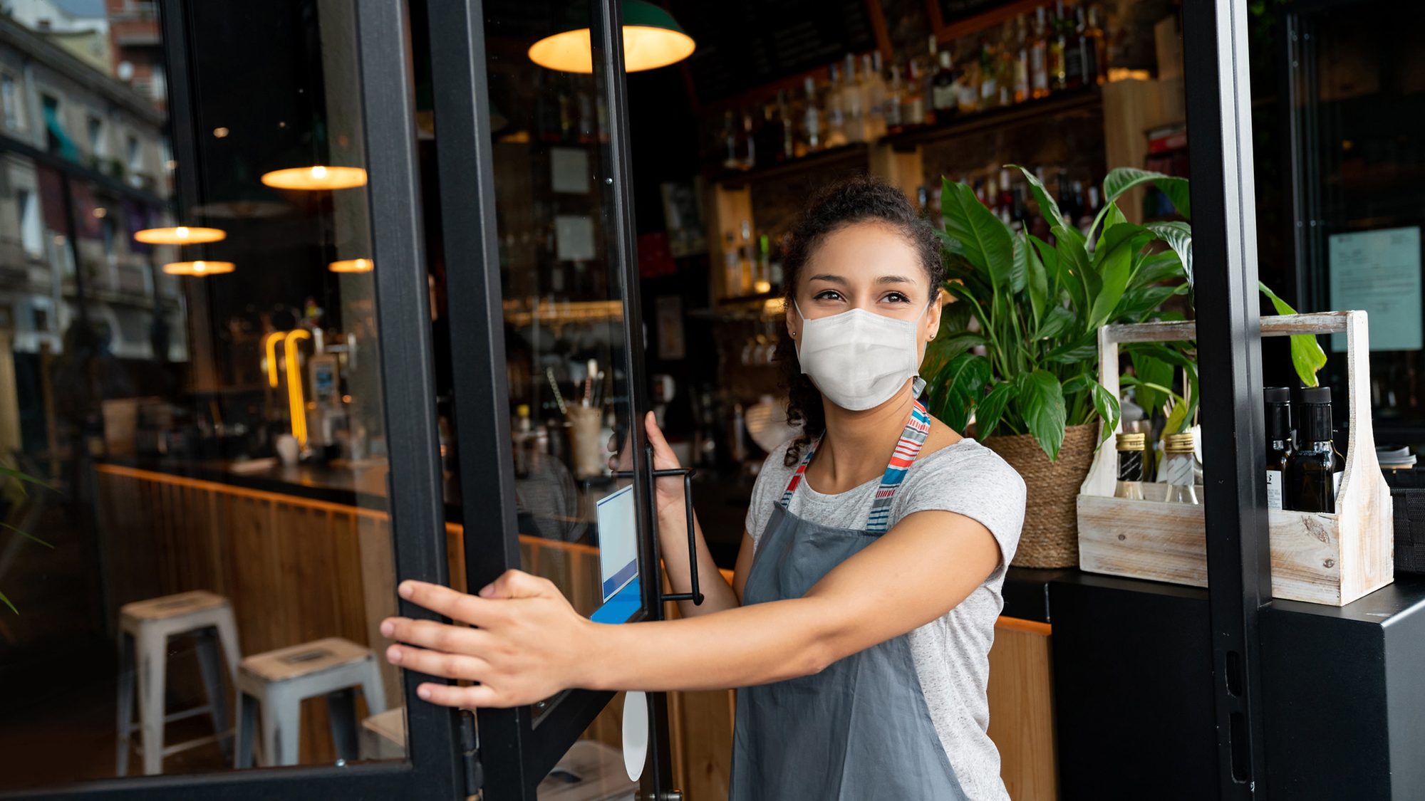 Woman in mask and apron, opening door to restaurant with plant in background.