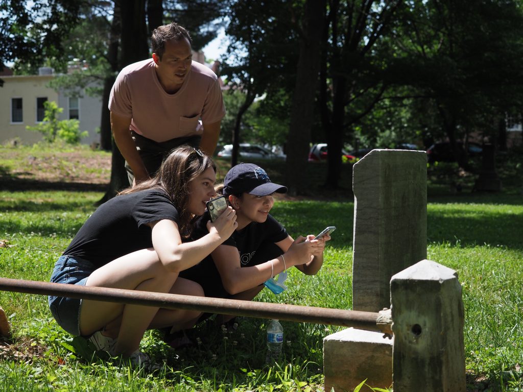 Man and two women point to inscriptions on a headstone