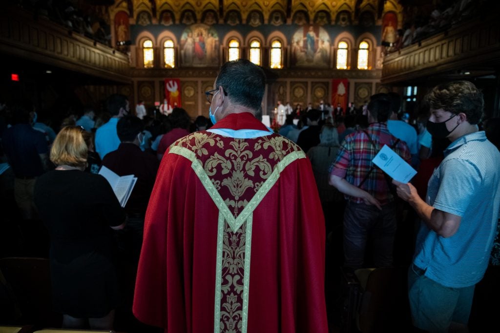 Back of a priest wearing a red robe walking through a full student audience