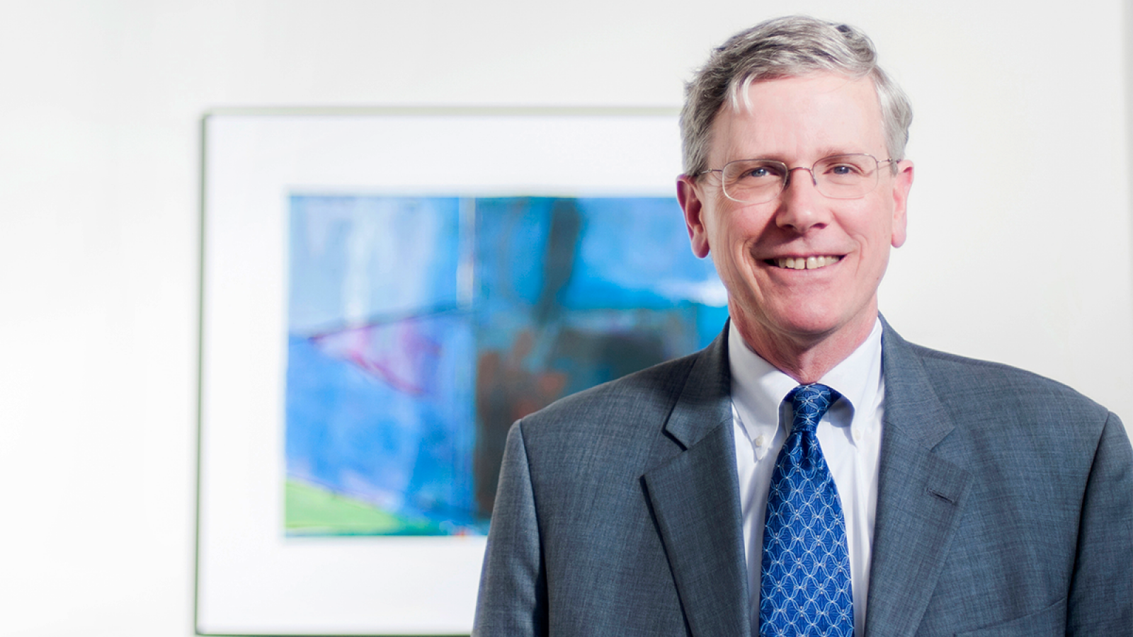 John Monahan wears a suit in front of a blue picture on white wall