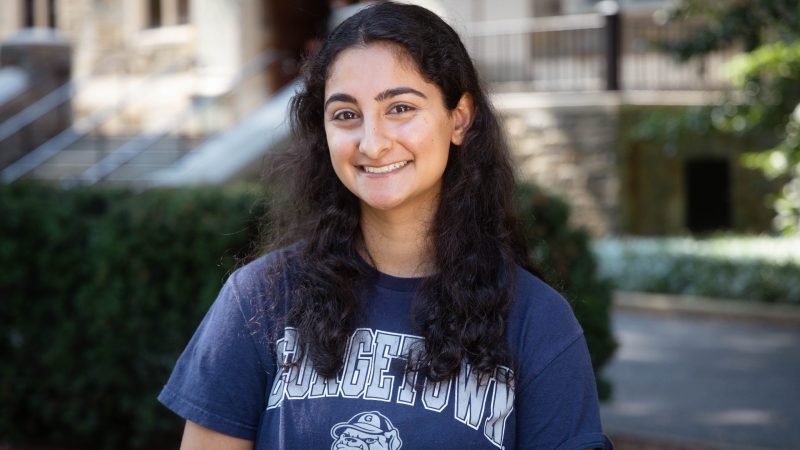 Zehra Mirza wears a dark blue Georgetown t-shirt in front of a stone building and bush