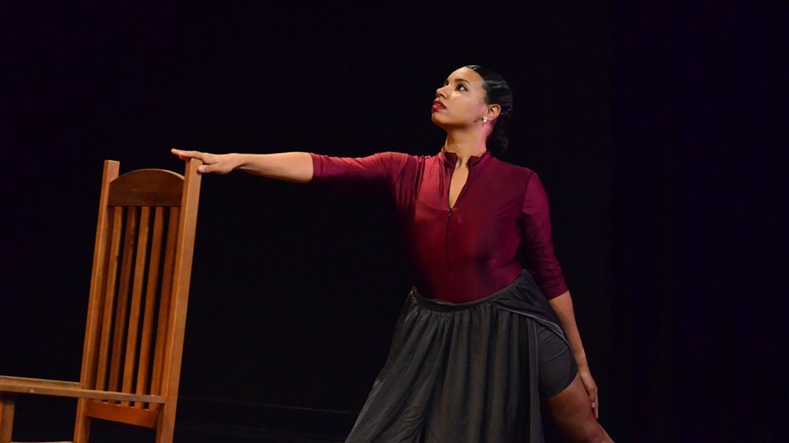 Prof. Raina Lucas in a burgundy top and gray skirt lunges behind a brown chair with her arm outstretched, against a black background.