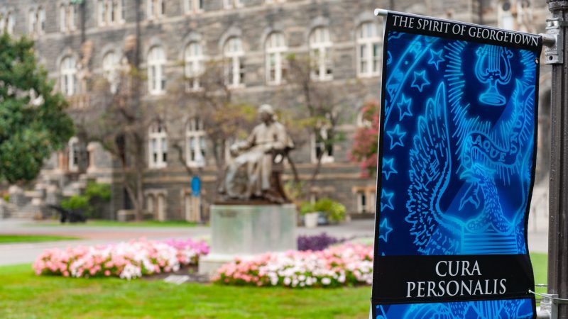 Spirit of Georgetown sign with light blue seal and the text &quot;Cura Personalis&quot; in front of a statue of John Carroll and Healy Hall