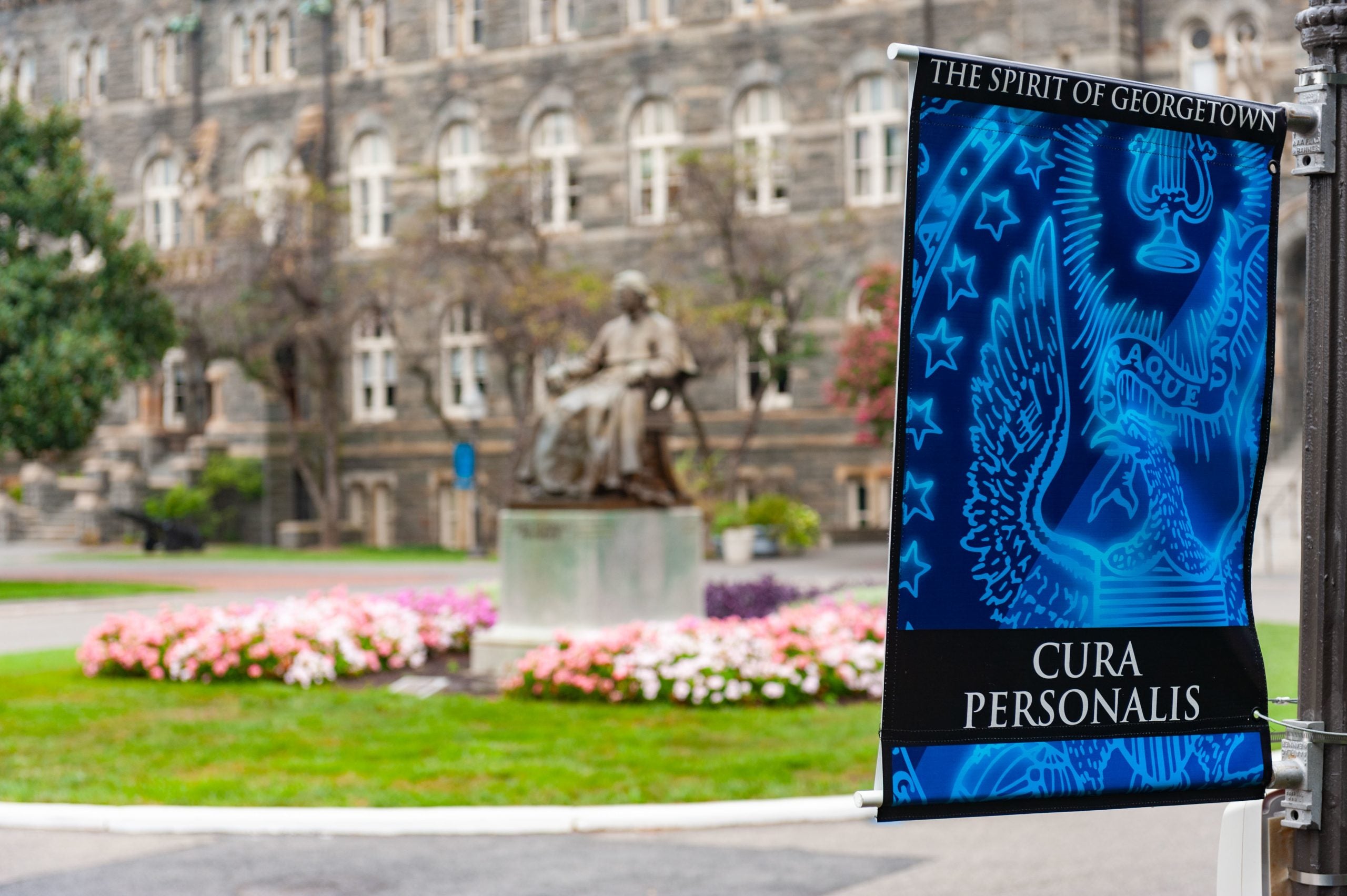 Spirit of Georgetown sign with light blue seal and the text "Cura Personalis" in front of a statue of John Carroll and Healy Hall