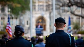 A female and male member of the military stand in uniform facing White-Gravenor Hall with an American flag in the foreground.