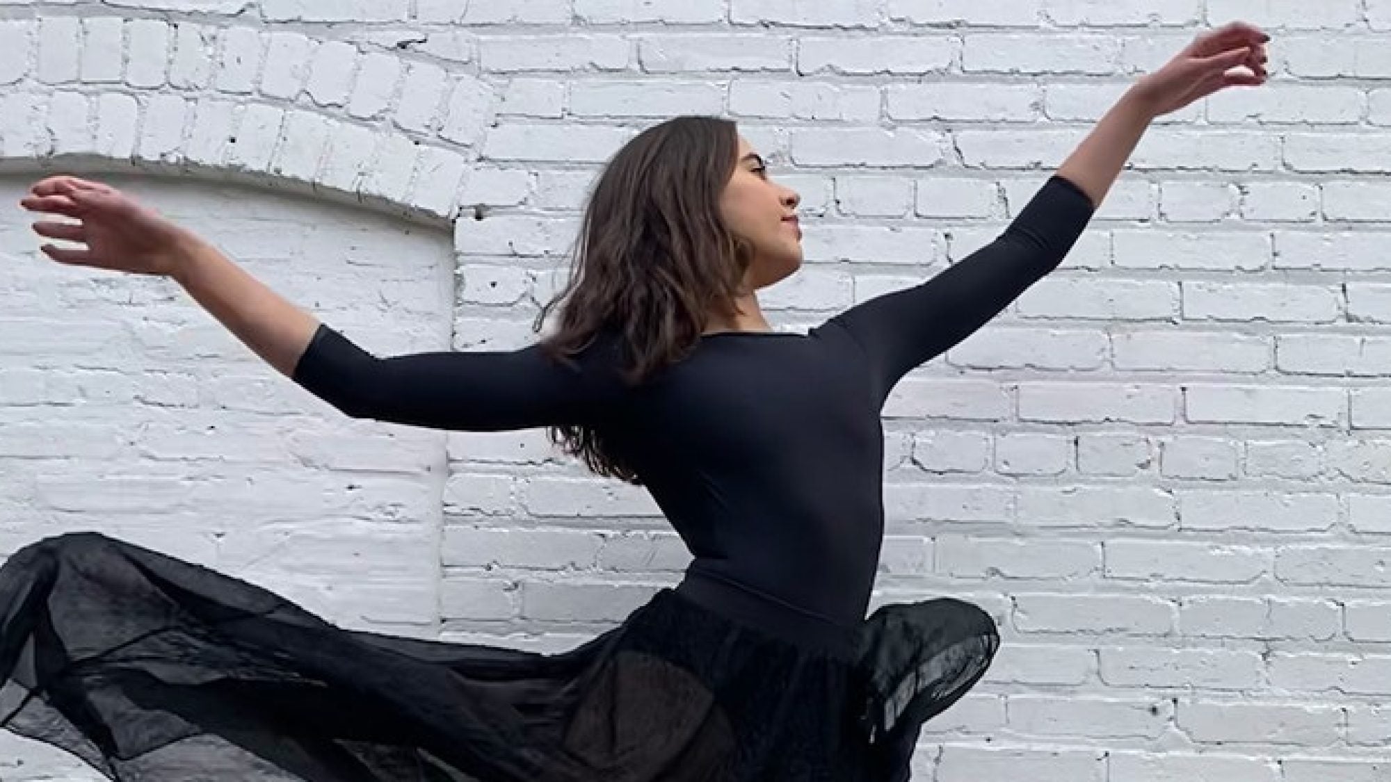 A Georgetown University Dance Company student in a black leotard and skirt raises her arms, against a white brick wall background.