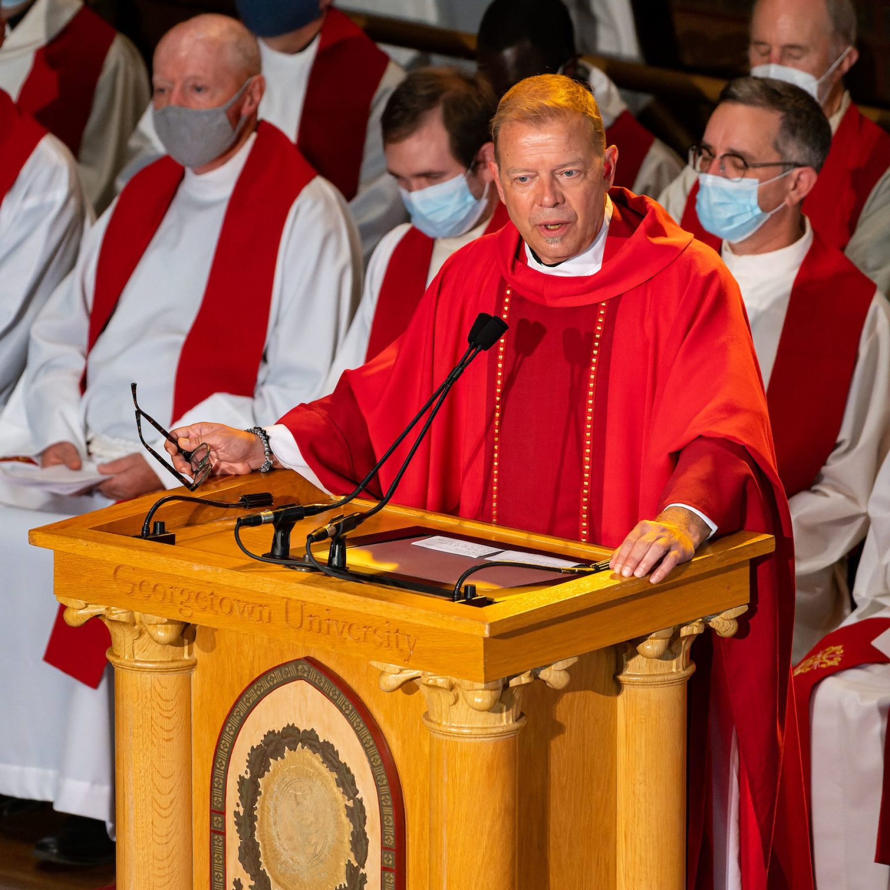 Fr. Greg Schenden, S.J., wears red robes while speaking at a podium