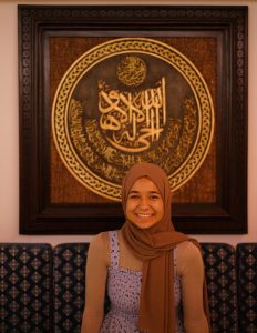 Doha Maaty stands in front of a an ornate framed prayer