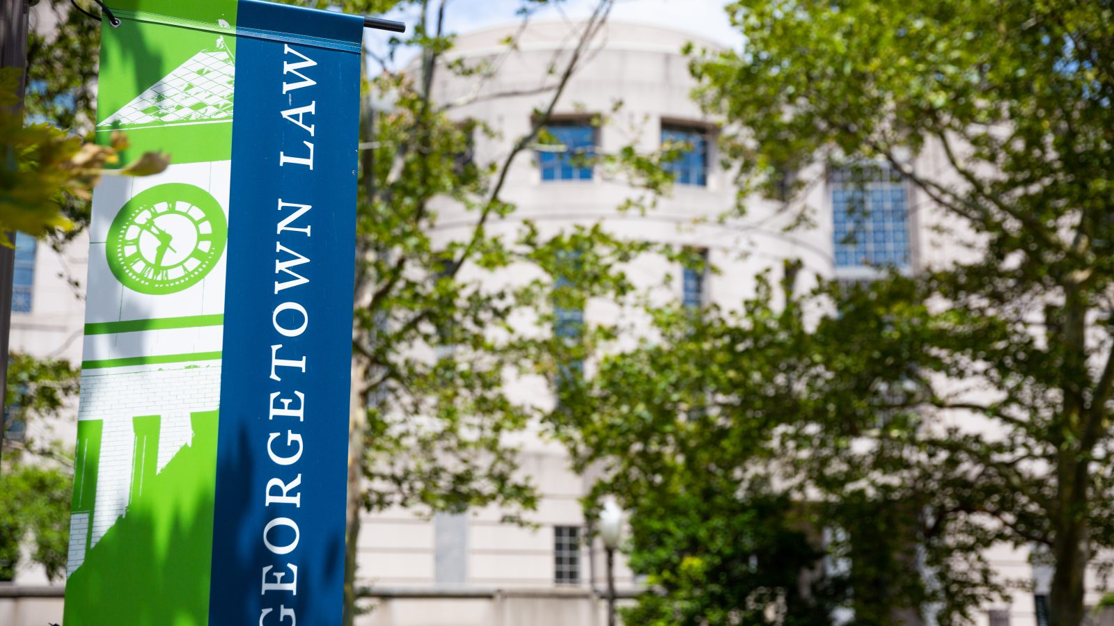 An image of a blue flag (left) with the words &quot;Georgetown Law&quot; in front of a white Georgetown Law building with green trees surrounding it.