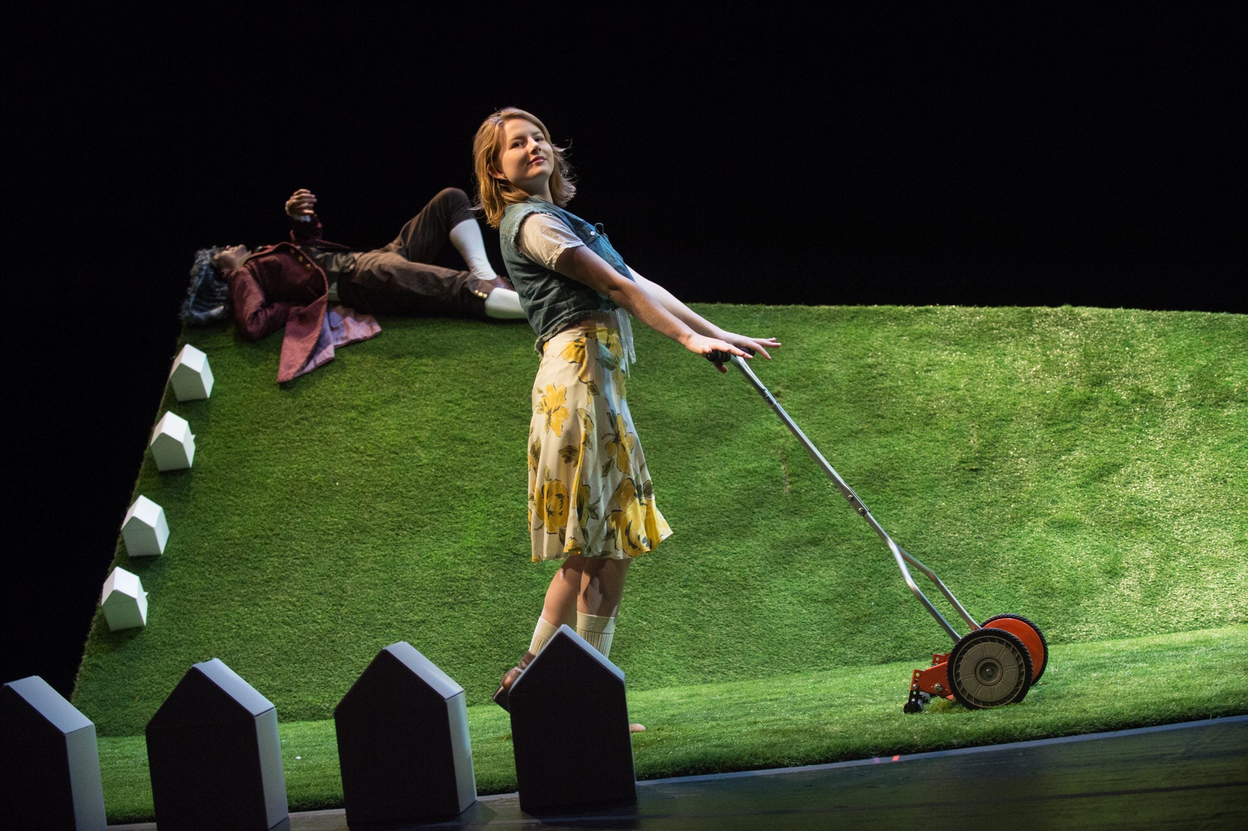 Actor proudly holds a lawn mower on a fake grass set onstage