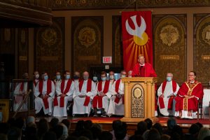 Priest in red robes delivers a speech from a podium in front of other men in white and red robes