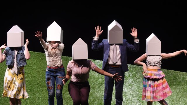 Student actors with houses covering their heads on a grass set