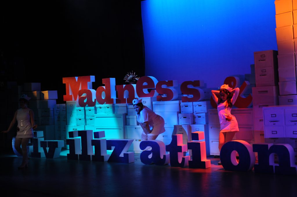 Actors dressed as nurses stand around large letters spelling out "Madness & Civilization"