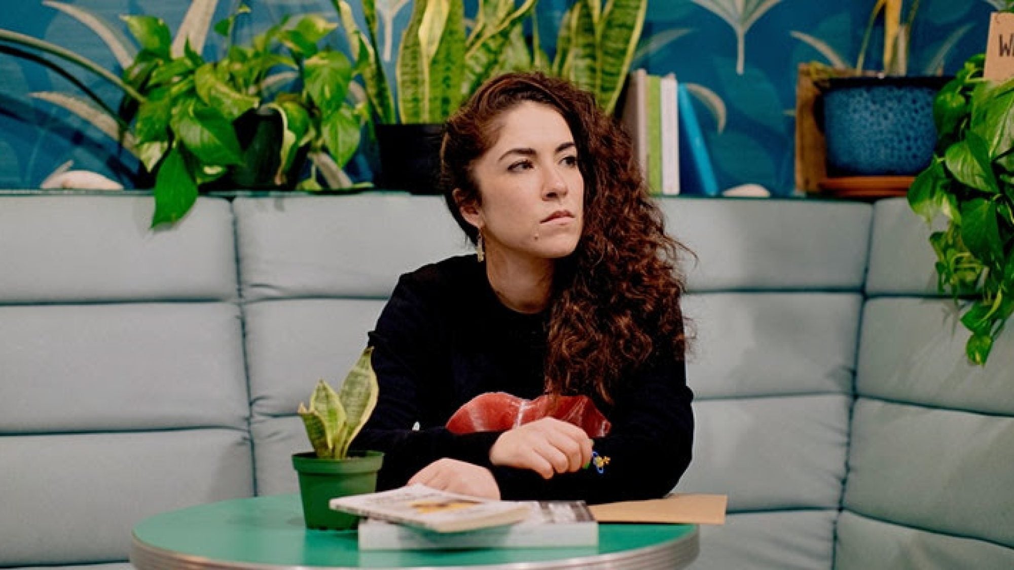 Georgetown University alumna Caitlin Cassidy sits at a green bistro table, looking off into the distance and surrounded by a shelf of houseplants and books.