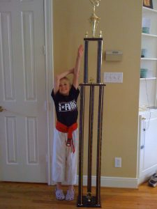 A young Jordan Kramer wears taekwondo cloths next to a trophy taller than she is