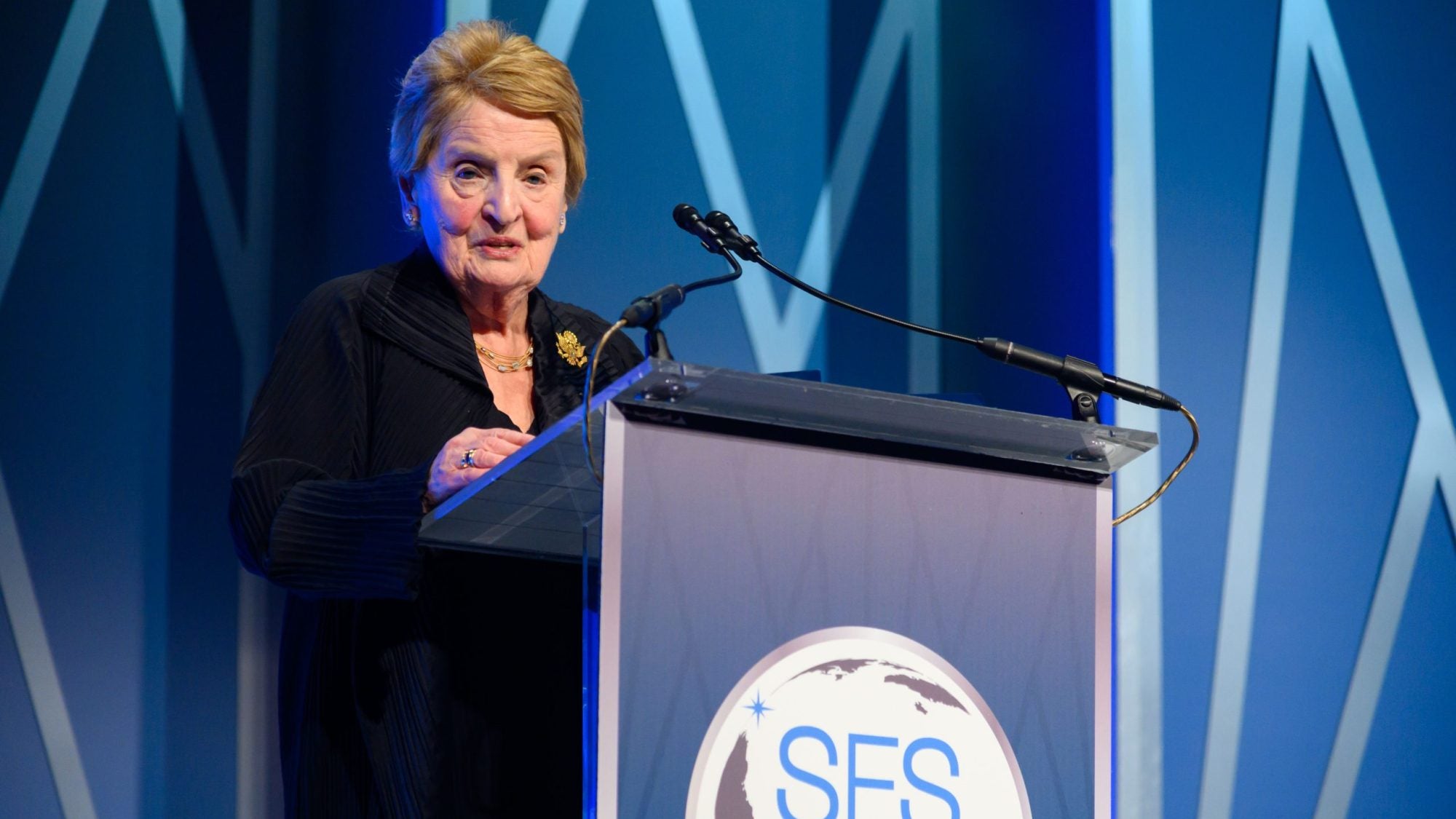 Madeleine Albright stands behind a podium with a blue background