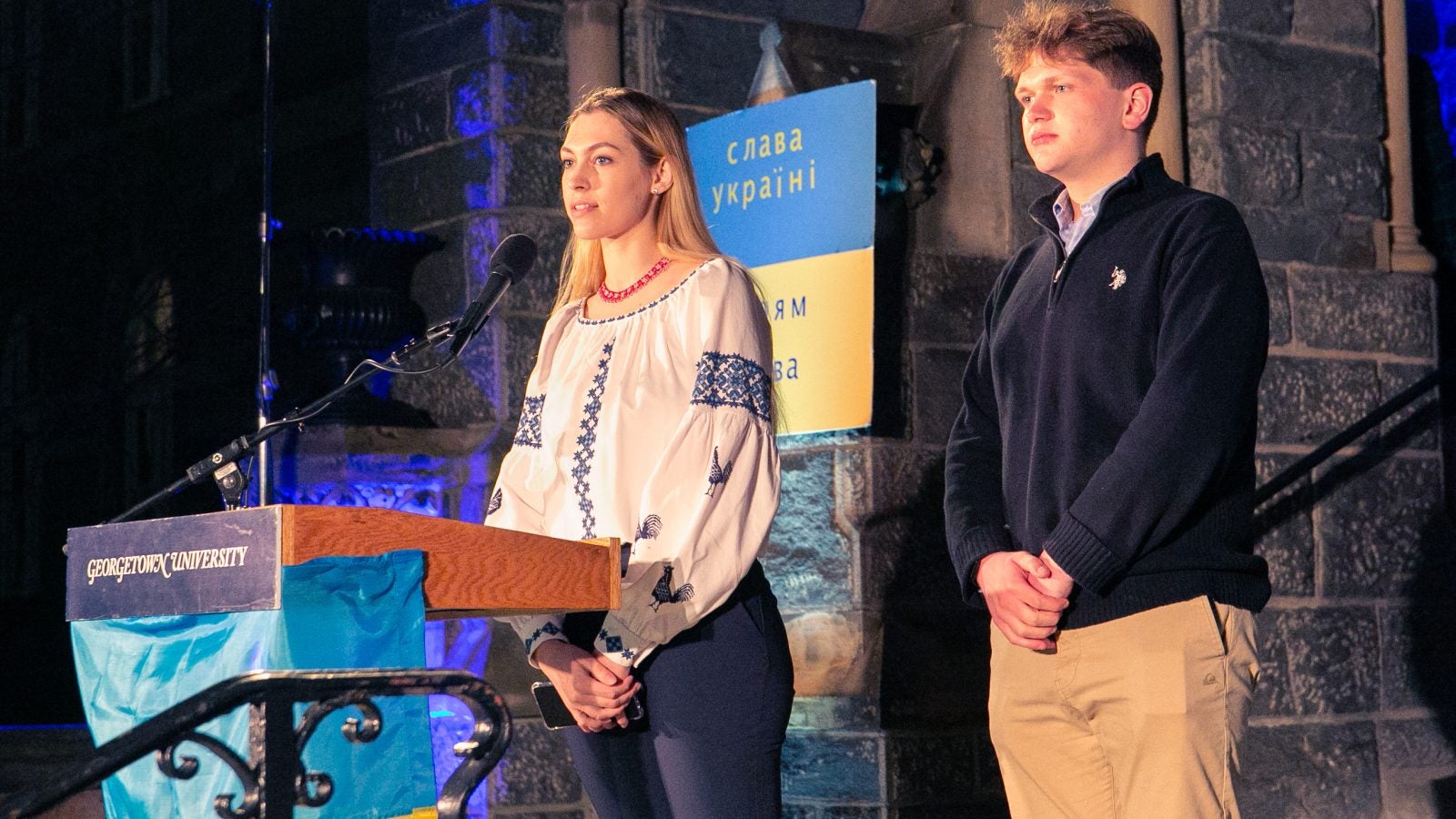 Two students speak from behind a podium with the Ukrainian flag across the front