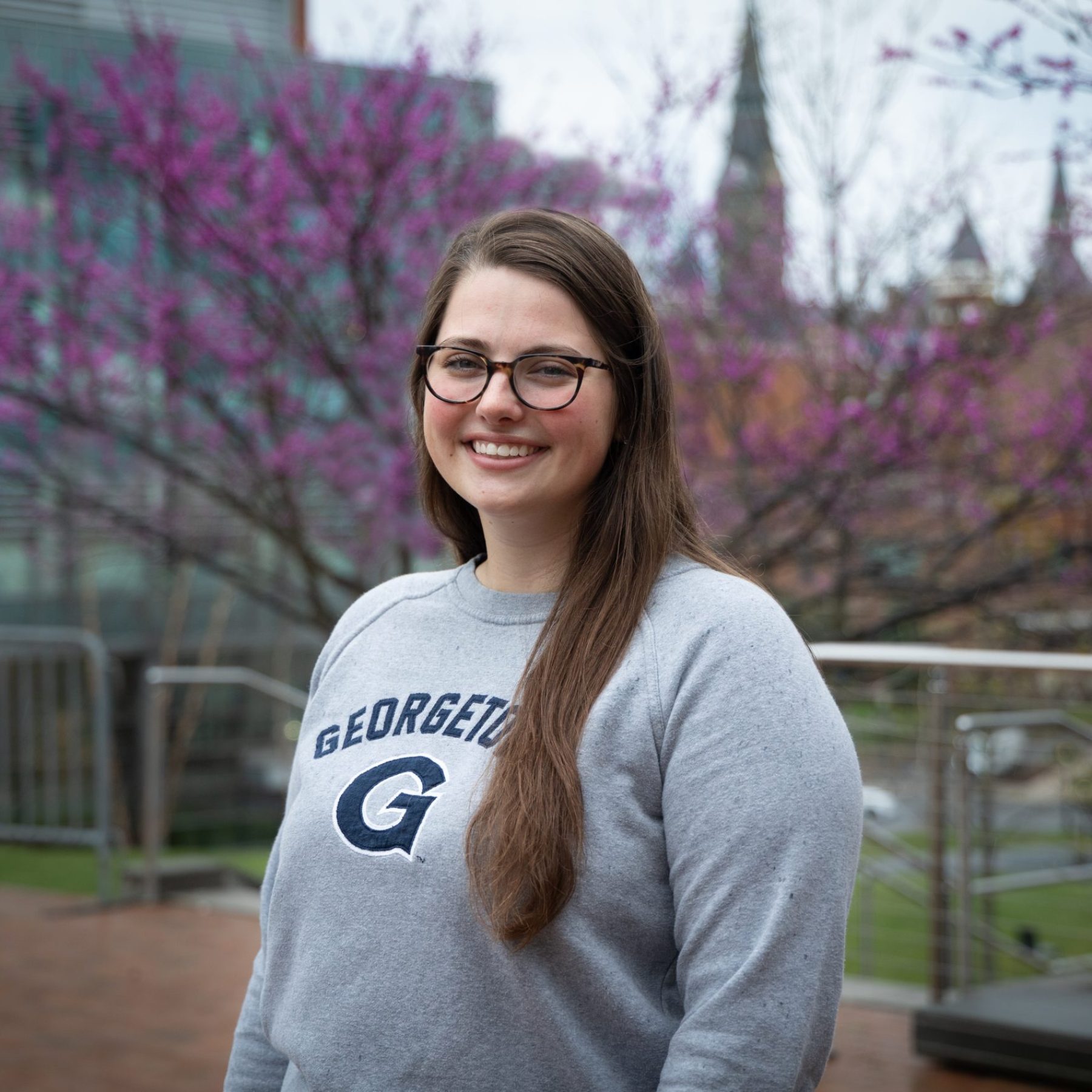 Kiki wears a gray Georgetown sweatshirt while outside in front of a blooming tree with Healy Hall in the background