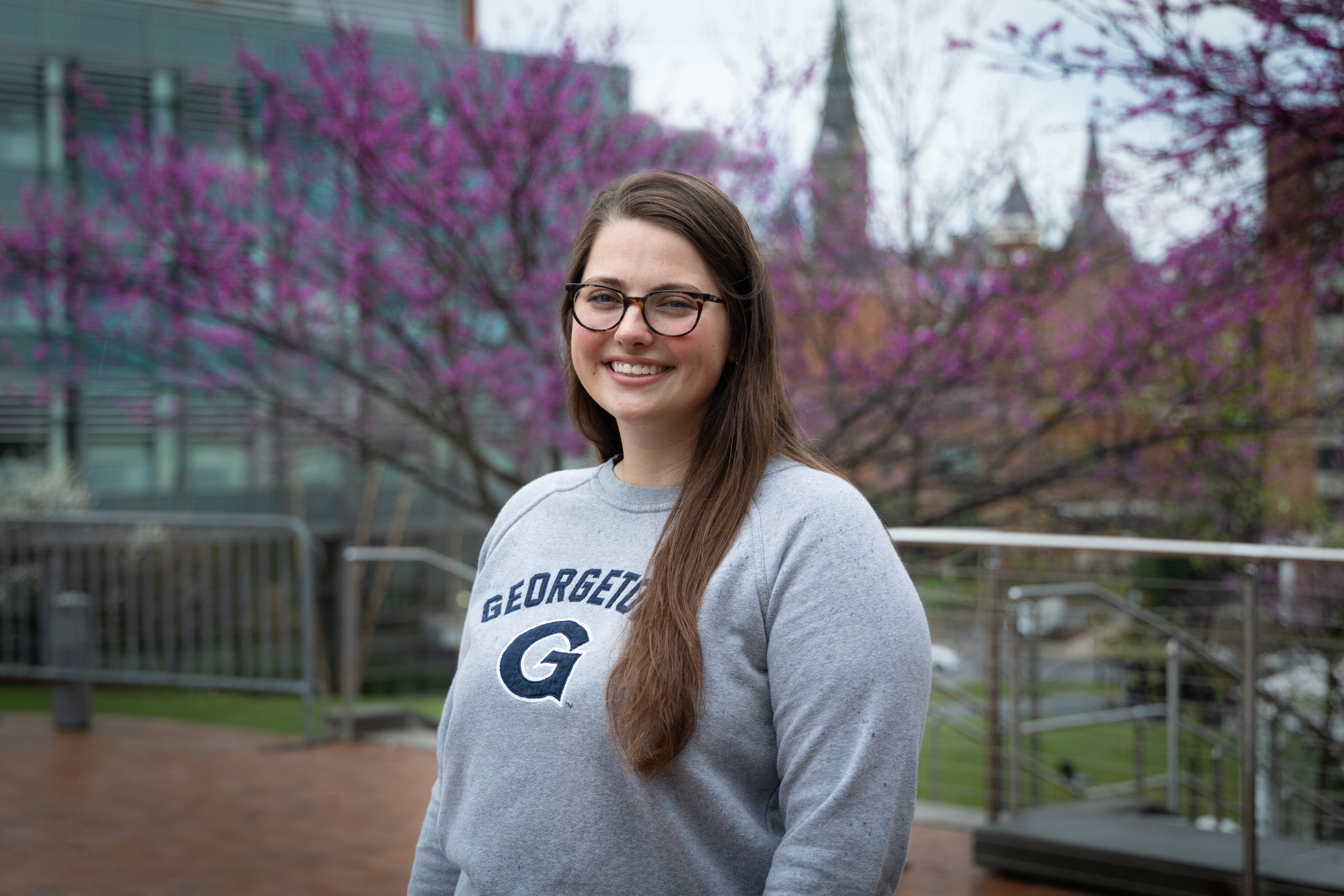 Kiki wears a gray Georgetown sweatshirt while outside in front of a blooming tree with Healy Hall in the background