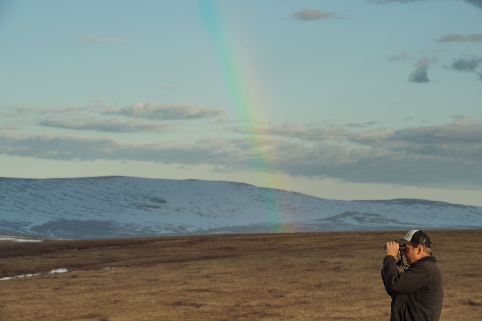 Pete Marra stands with binoculars with a rainbow in the background