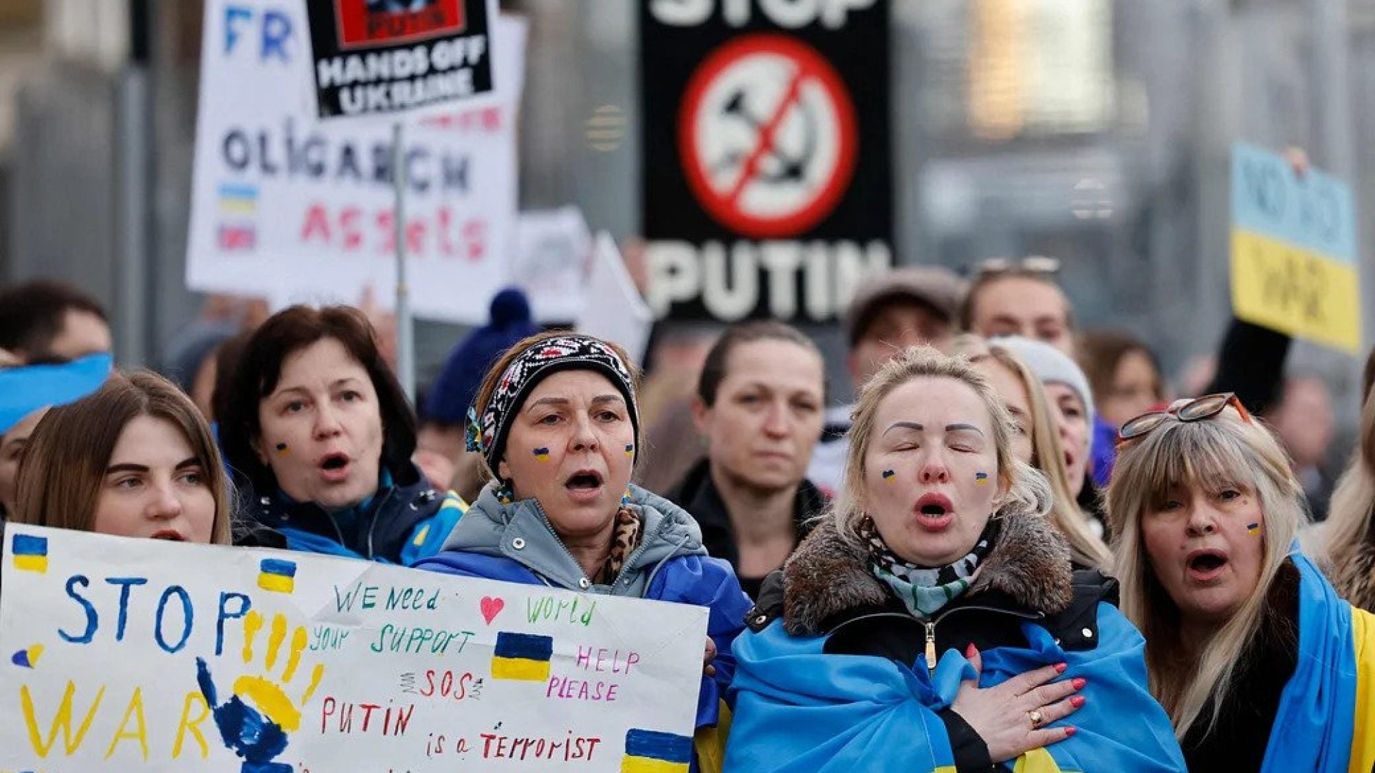 Women protesting, wrapped in Ukraine flags, holding signs.