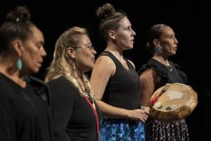 Four women perform onstage while one of them beats a drum