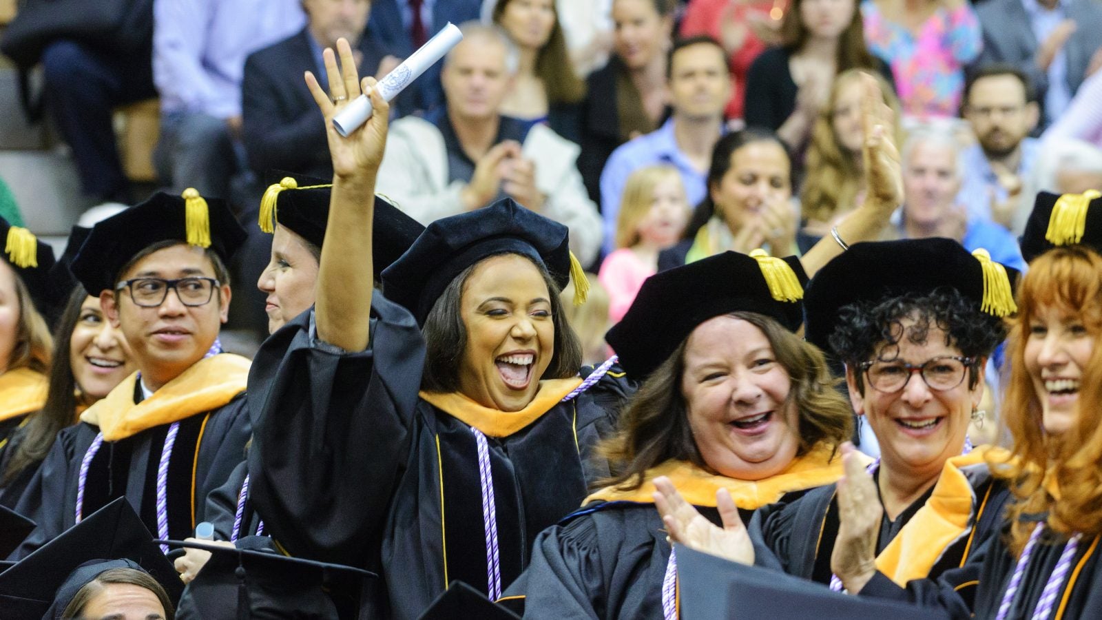 Student wearing graduation attire holding a diploma cheers in a crowd