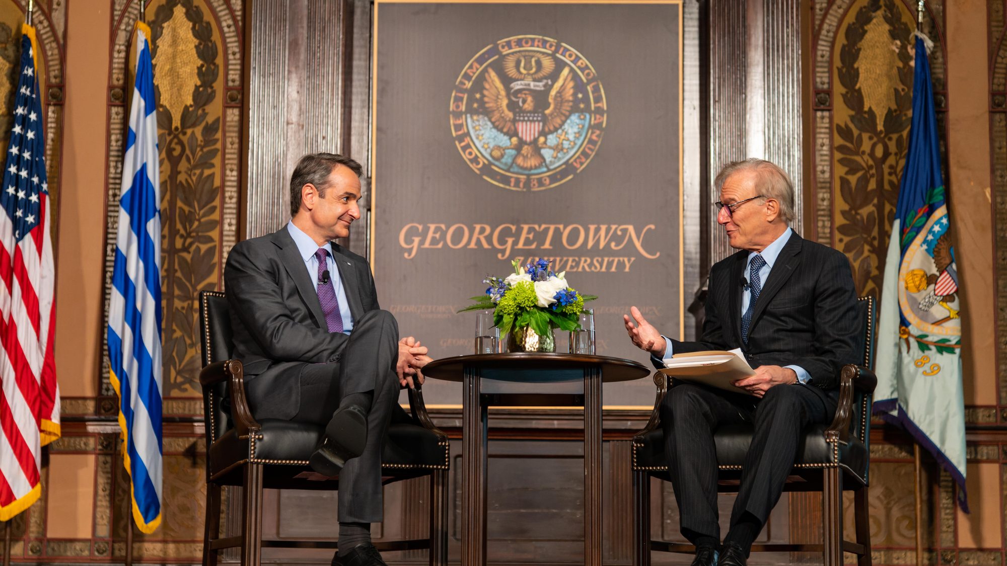 Two men in suits speak onstage from either end of a table with flowers on it with in front of a Georgetown University sign