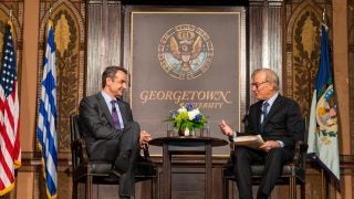 Two men in suits speak onstage from either end of a table with flowers on it with in front of a Georgetown University sign