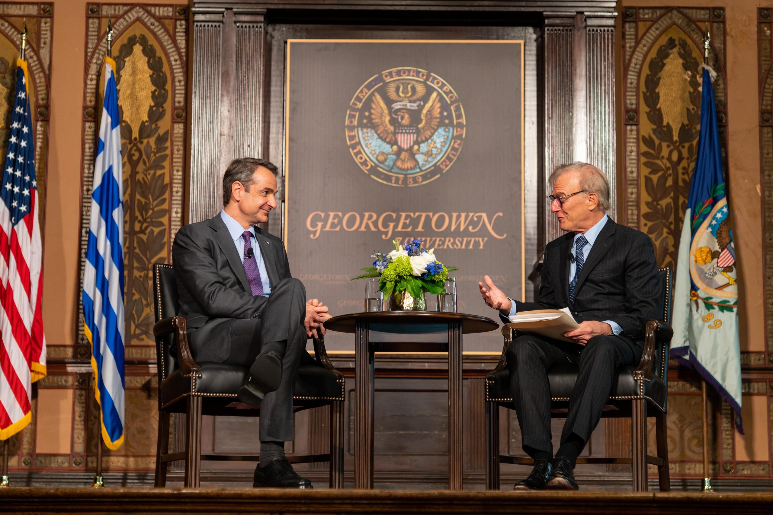 Two men in suits speak onstage from either end of a table with flowers on it with in front of a Georgetown University sign