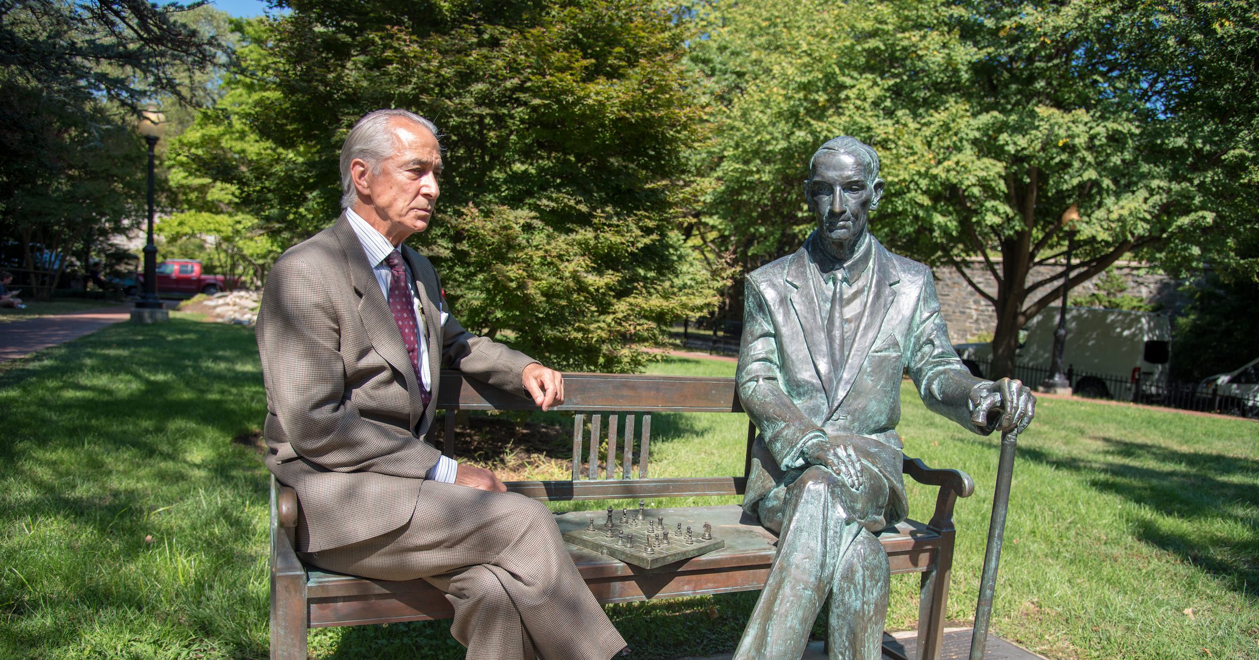 Strathairn as Karski sits on a bench next to a greening Karski statue with a cane