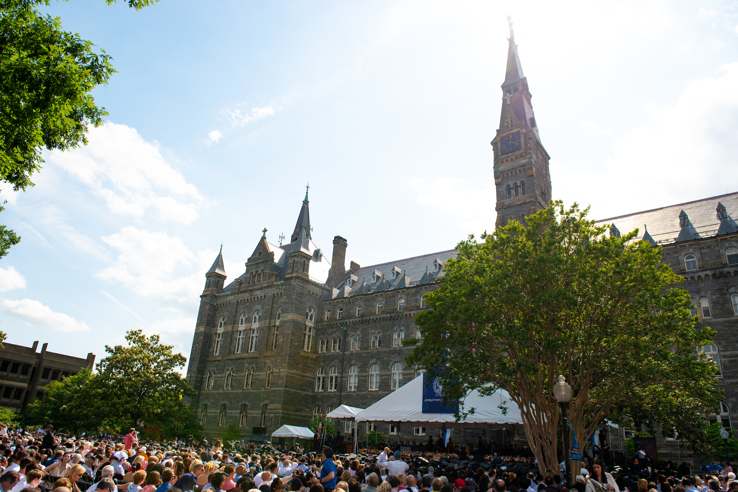 Commencement crowd in seats in front of Healy Hall with the sun shining behind the clock tower