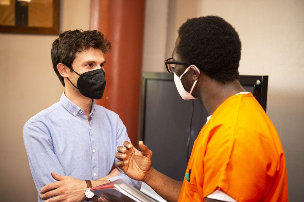 Omar Alshogre (left) wears a mask and a blue button down and talks with a scholar in Georgetown's Prison Scholars Program who wears an orange jumpsuit and a mask.