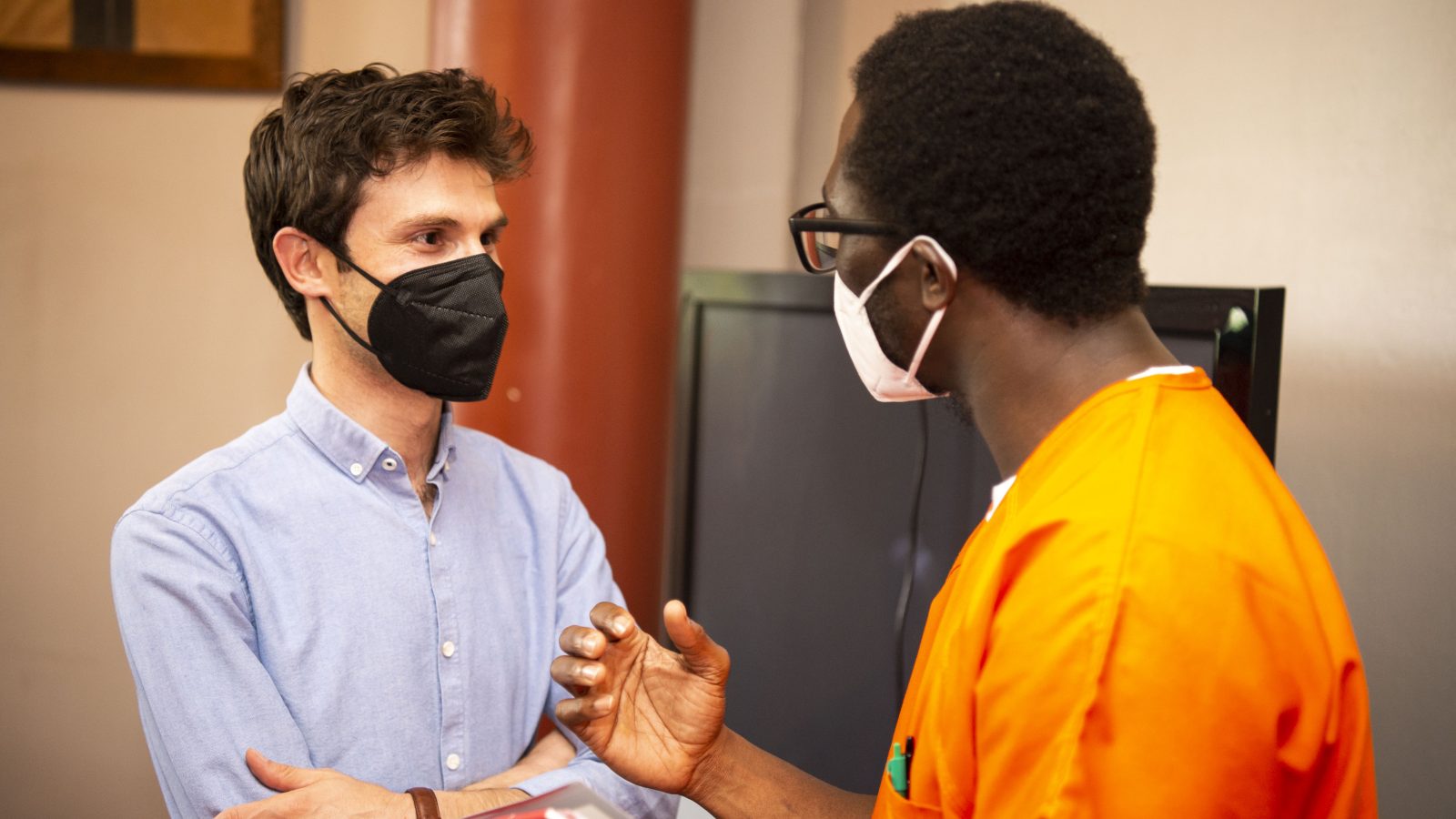 Omar Alshogre (left) wears a mask and a blue button down and talks with a scholar in Georgetown&#039;s Prison Scholars Program who wears an orange jumpsuit and a mask.