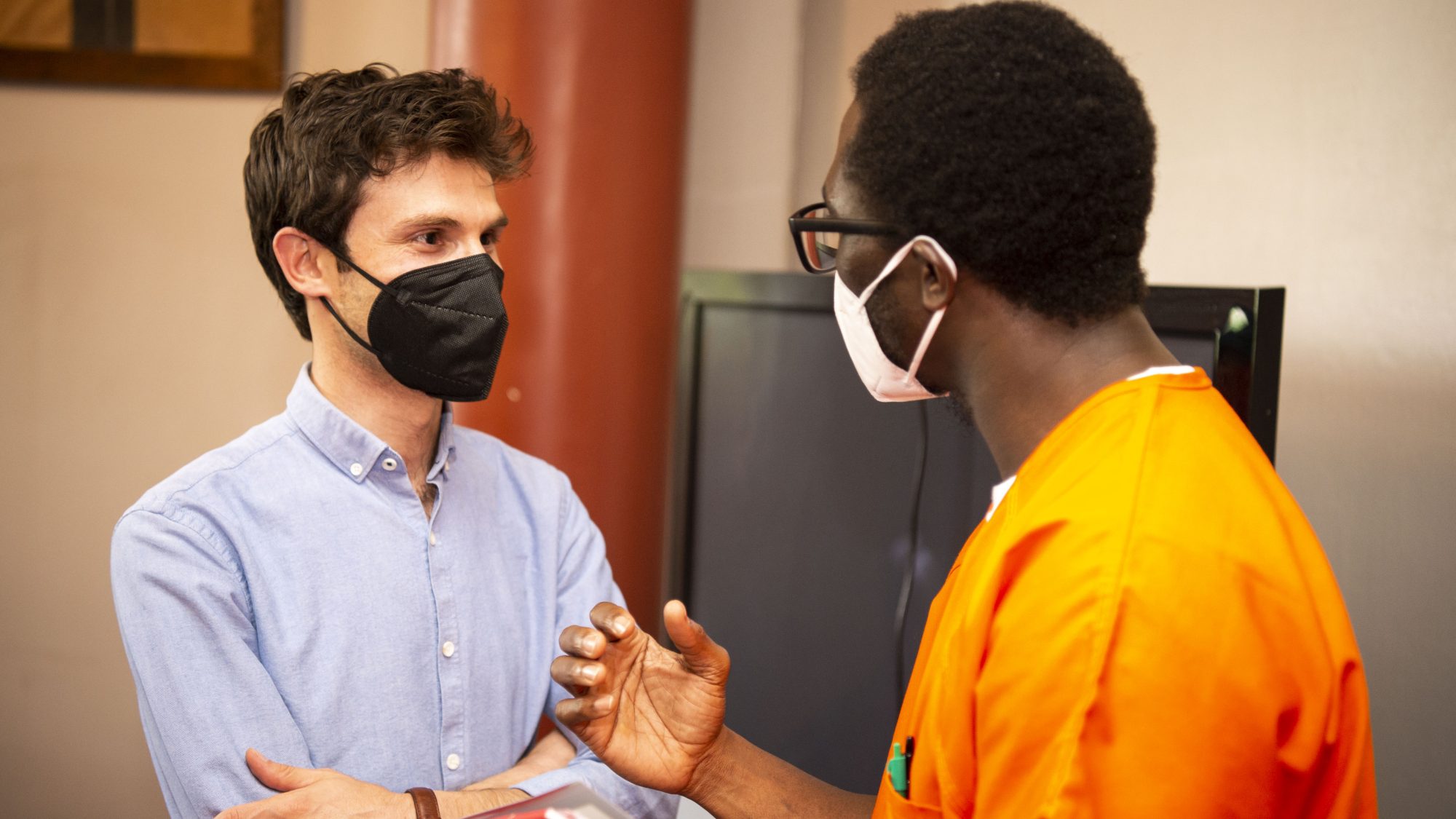 Omar Alshogre (left) wears a mask and a blue button down and talks with a scholar in Georgetown's Prison Scholars Program who wears an orange jumpsuit and a mask.