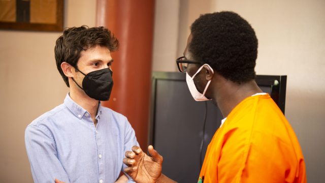 Omar Alshogre (left) wears a mask and a blue button down and talks with a scholar in Georgetown&#039;s Prison Scholars Program who wears an orange jumpsuit and a mask.