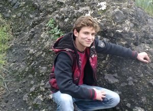 Omar, pictured at age 15, poses in front of a rock in Syria wearing a red shirt and black jacket