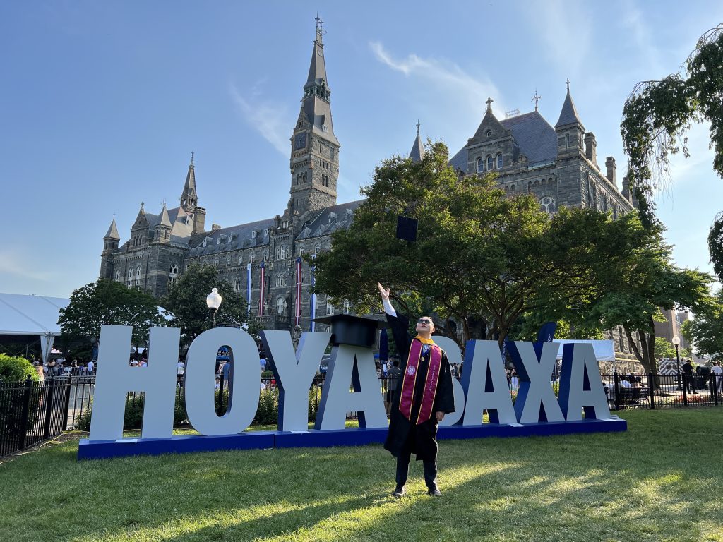 Charlie Wang throws his graduation hat in the air in front of a sign that says "Hoya Saxa" on Healy Lawn.